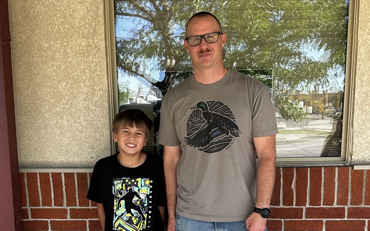 A Big and Little standing together in front of a brick wall and window